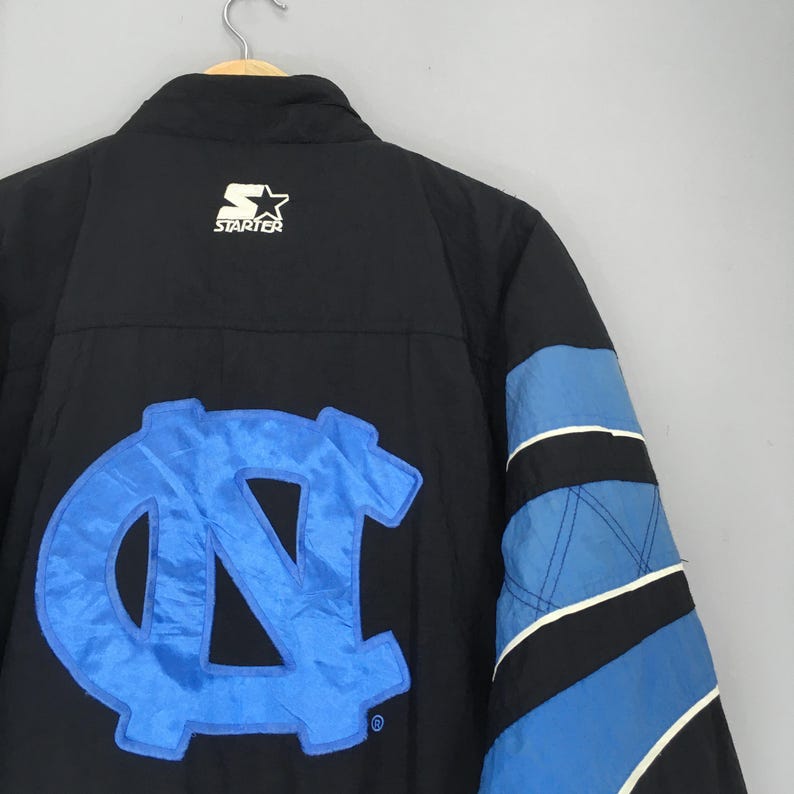 May include: Black jacket with a large light blue "NC" logo on the back. The jacket has a Starter logo on the upper back and blue and black panels on the sleeve. The jacket is hanging on a wooden hanger.
