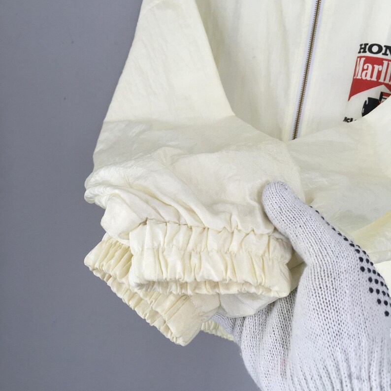 May include: A cream-colored windbreaker jacket featuring a full-length front zipper and elasticized cuffs. The chest area displays a graphic logo with the text Honda and Marlboro in red, black, and white colors on a textured fabric surface.