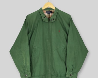 May include: A forest green zip-up jacket featuring a collared neckline, long sleeves with buttoned cuffs, and two front pockets. The interior lining displays a tan, black, and red plaid pattern. A small red embroidered logo is on the left chest.