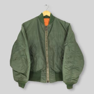 May include: An olive green bomber jacket with a full zip, ribbed cuffs, and a collar. The jacket has an orange lining and is suspended on a wooden hanger. It features two front pockets and a classic bomber design. The jacket is a classic style.