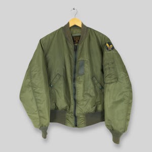 Può includere: Giacca bomber verde oliva con zip, polsini e orlo a coste. La giacca presenta una toppa sulla manica superiore sinistra e una piccola tasca sulla manica sinistra. Appesa a una gruccia di legno.