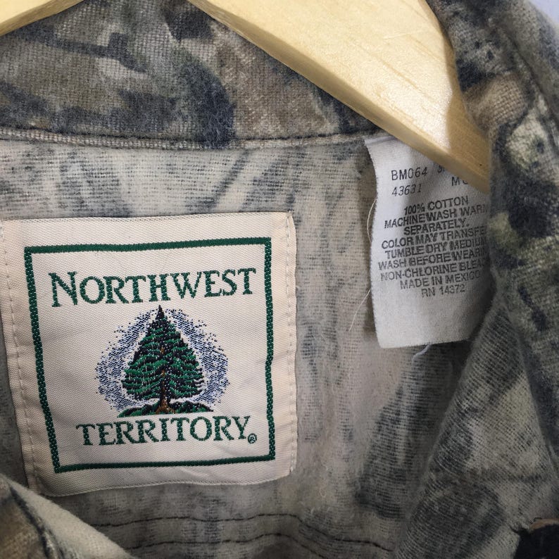 May include: Close-up of a camouflage-patterned shirt with a Northwest Territory label. The label features a green-bordered rectangle with the brand name and a pine tree graphic. A tag with washing instructions is also visible.