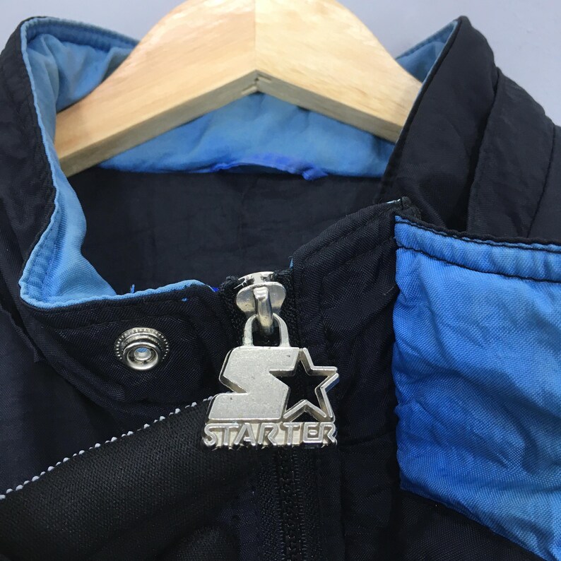 May include: Close-up of a vintage black and blue Starter jacket. The jacket features a silver zipper pull with the Starter logo and a snap closure. The collar and accents are light blue.