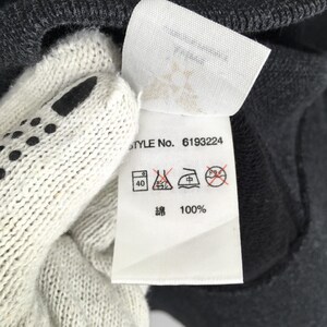 Pu&ograve; includere: Primo piano di un'etichetta bianca per abbigliamento con il testo "STYLE No. 6193224" e simboli di cura. L'etichetta include anche il testo "綿 100%" ed &egrave; parzialmente oscurata da un capo grigio e da un guanto bianco.