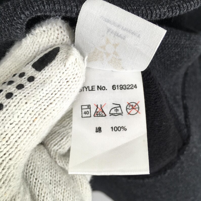 Pu&ograve; includere: Primo piano di un'etichetta bianca per abbigliamento con il testo "STYLE No. 6193224" e simboli di cura. L'etichetta include anche il testo "綿 100%" ed &egrave; parzialmente oscurata da un capo grigio e da un guanto bianco.
