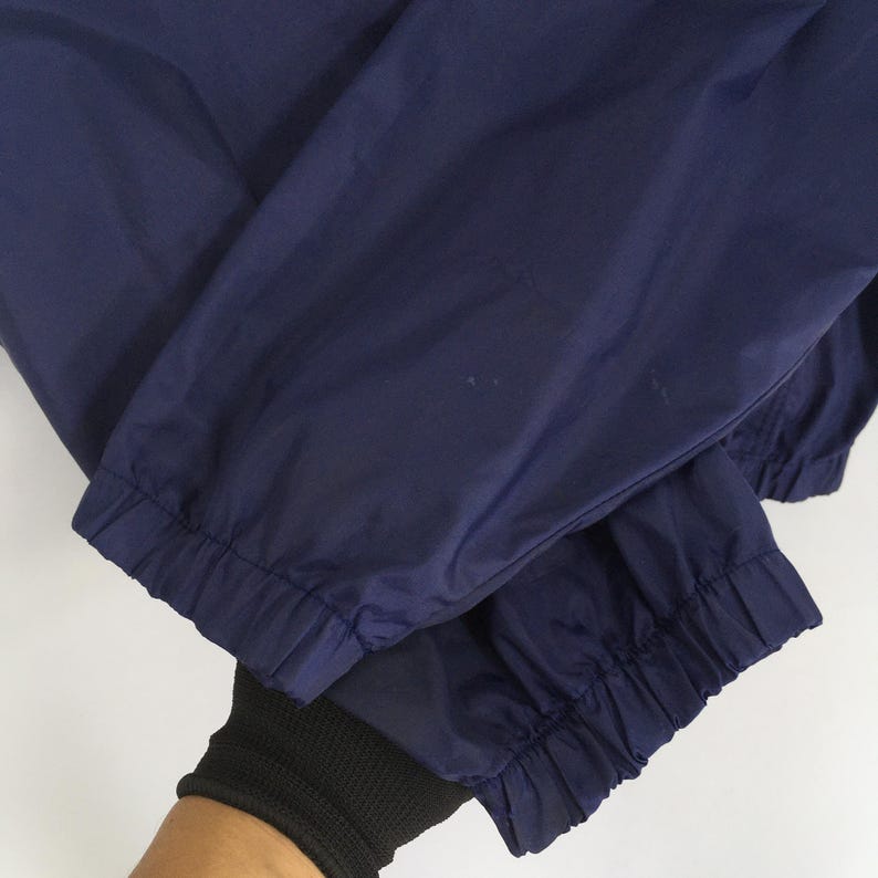 Pu&ograve; includere: Primo piano di pantaloni sportivi blu navy con polsini elasticizzati. I pantaloni sono realizzati in un materiale leggero, forse nylon. I polsini sono raccolti con elastico. I pantaloni sono piegati.
