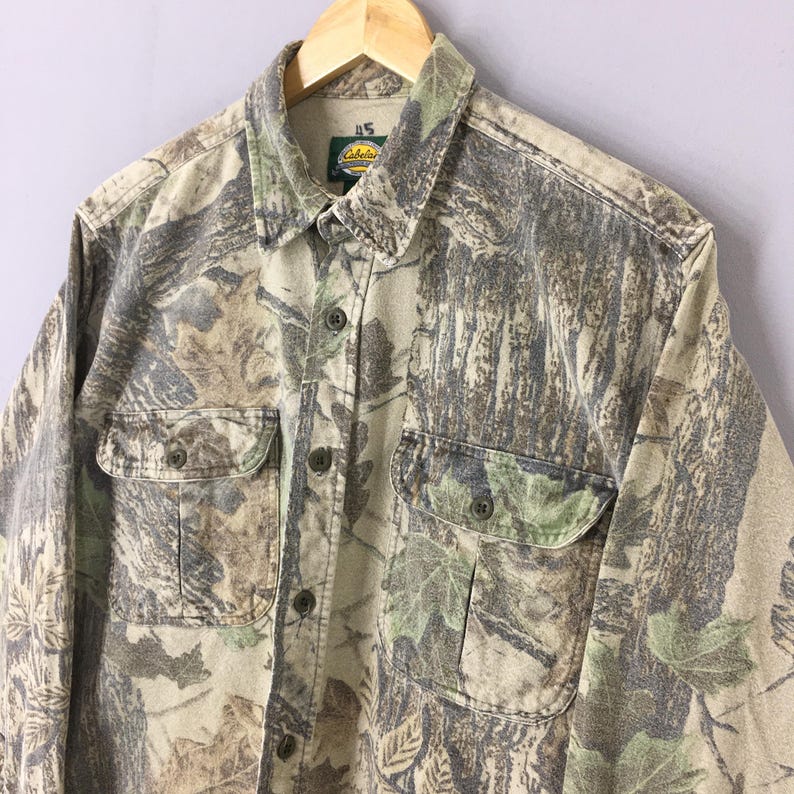 Puede incluir: Una camisa de camuflaje de manga larga con dos bolsillos en el pecho y botones. El patr&oacute;n de bosque presenta tonos verdes, marrones y beige. La etiqueta de la marca Cabela's es visible en el cuello.