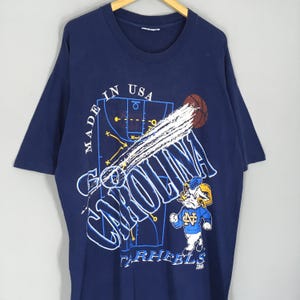 May include: Navy blue t-shirt with a basketball-themed graphic. The design includes the words "Go Carolina" in white, a basketball, and a cartoon ram mascot wearing a blue jersey. The shirt also has the text "Made in USA".