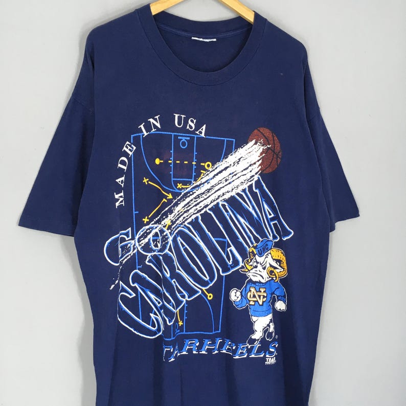 May include: Navy blue t-shirt with a basketball-themed graphic. The design includes the words "Go Carolina" in white, a basketball, and a cartoon ram mascot wearing a blue jersey. The shirt also has the text "Made in USA".
