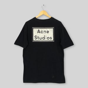 May include: Black t-shirt with the text "Acne Studios" in a white rectangle on the back. The text is in a bold, sans-serif font. The shirt is hanging on a wooden hanger.