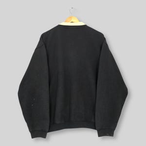 May include: Black sweatshirt with a cream-colored collar, hanging on a wooden hanger. The sweatshirt has long sleeves and a crew neck. The back of the sweatshirt is visible, showing a simple, casual design.