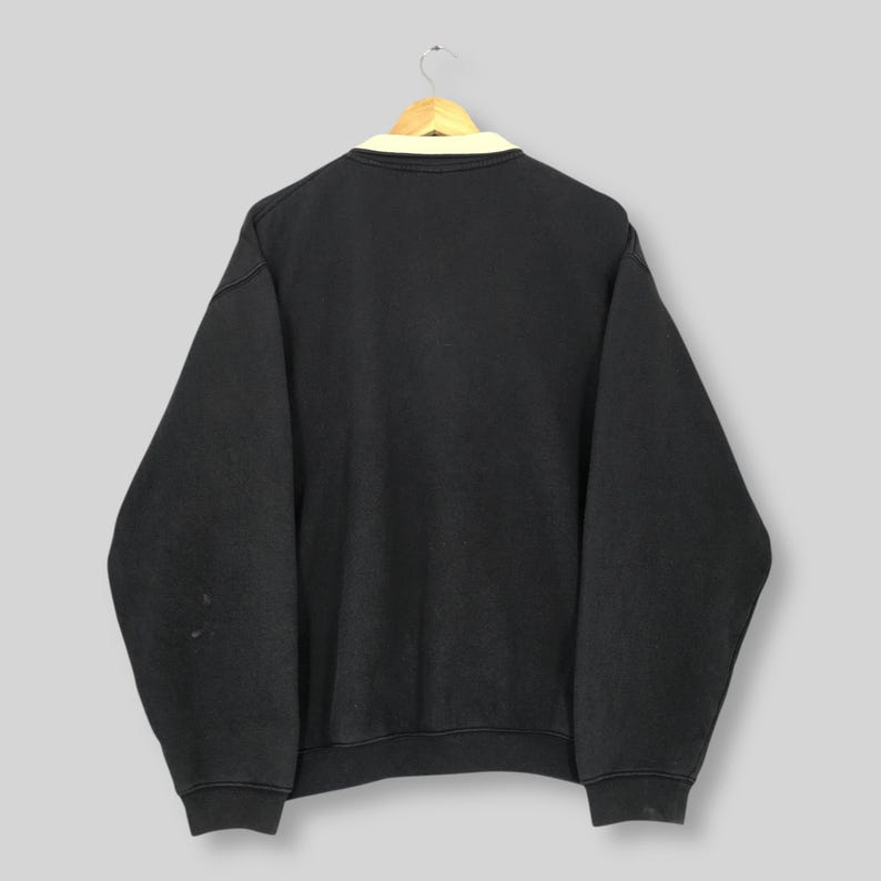 May include: Black sweatshirt with a cream-colored collar, hanging on a wooden hanger. The sweatshirt has long sleeves and a crew neck. The back of the sweatshirt is visible, showing a simple, casual design.