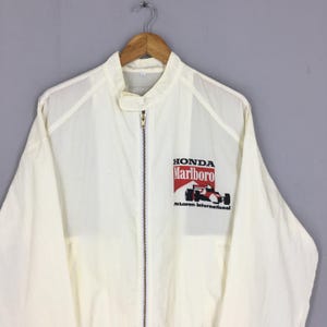 May include: A white long-sleeve zip-up jacket featuring a stand-up collar with a snap closure. The chest displays a graphic with the text Honda, Marlboro, and McLaren International, accompanied by an illustration of a Formula One race car.