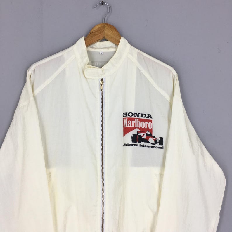 May include: A white long-sleeve zip-up jacket featuring a stand-up collar with a snap closure. The chest displays a graphic with the text Honda, Marlboro, and McLaren International, accompanied by an illustration of a Formula One race car.