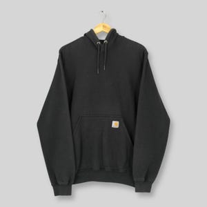 May include: A charcoal gray hoodie with a drawstring hood and a kangaroo pocket. The hoodie features a small, square logo patch on the pocket. The garment is hanging on a wooden hanger.