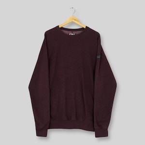 May include: A maroon crew neck sweatshirt with long sleeves hangs from a wooden hanger. The sweatshirt has a textured appearance and a small black label on the left sleeve. The background is a solid light gray.