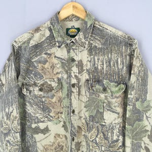 Puede incluir: Camisa con botones de camuflaje con dos bolsillos en el pecho. La camisa presenta un patr&oacute;n de camuflaje de bosque en tonos verdes, marrones y beige. La camisa tiene cuello y est&aacute; colgada en una percha de madera.