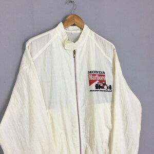 May include: A cream-colored lightweight windbreaker jacket featuring a full-length front zipper, a high collar with a tab closure, and a graphic on the chest displaying the text Honda, Marlboro, and McLaren International with a race car illustration.
