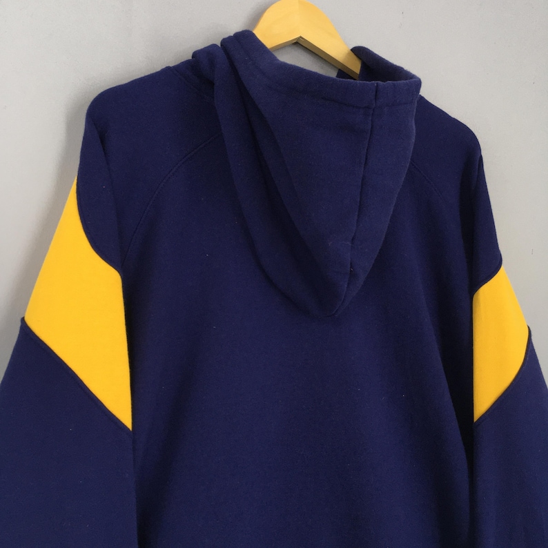 May include: A navy blue hoodie with yellow shoulder panels. The garment is suspended on a wooden hanger against a light grey backdrop. The rear view of the sweatshirt displays the hood and the colour-blocked design.