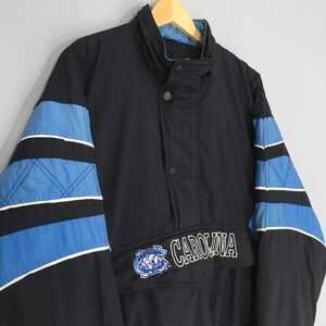 May include: Black jacket with blue and white accents on the sleeves. The jacket features the word "CAROLINA" in white lettering and a logo on the front. The jacket has a high collar and a button closure.