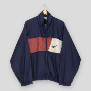 Pu&ograve; includere: Giacca a vento Nike blu navy con cerniera a tutta lunghezza. La giacca presenta una striscia rossa orizzontale sul petto, un pannello bianco con il logo Nike nero e un colletto rialzato. La giacca &egrave; appesa a una gruccia di legno.