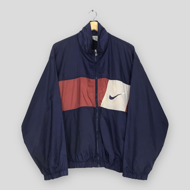 Pu&ograve; includere: Giacca a vento Nike blu navy con cerniera a tutta lunghezza. La giacca presenta una striscia rossa orizzontale sul petto, un pannello bianco con il logo Nike nero e un colletto rialzato. La giacca &egrave; appesa a una gruccia di legno.