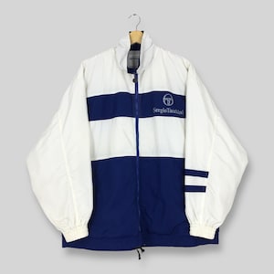 Op de afbeelding: Wit en marineblauw Sergio Tacchini trainingsjack. De jas heeft een ritssluiting over de hele lengte, een hoge kraag en het Sergio Tacchini-logo op de borst. Marineblauwe strepen lopen over de borst en mouwen.