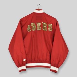 May include: A red varsity jacket featuring the text 49ERS embroidered in gold with white outlines across the back. The jacket includes a white collar, white cuffs, and a white waistband, with a small white Starter logo on the left sleeve cuff.