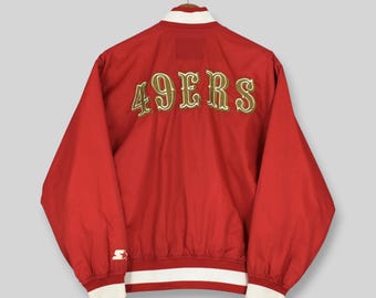 May include: A red varsity jacket featuring the text 49ERS embroidered in gold with white outlines across the back. The jacket includes a white collar, white cuffs, and a white waistband, with a small white Starter logo on the left sleeve cuff.