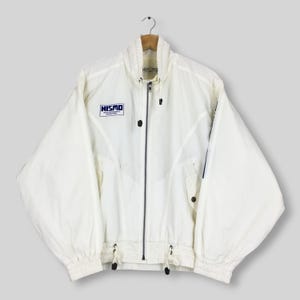 May include: A white windbreaker jacket featuring a high collar with a drawstring, a full-length front zipper, and long sleeves. The chest displays a blue NISMO logo. The jacket includes two front pockets with button closures and a drawstring hem.