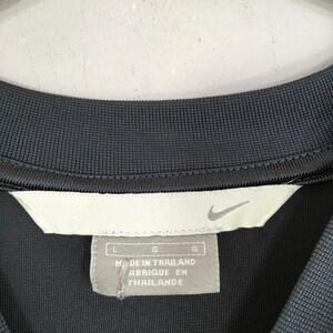 Puede incluir: Primer plano de un cuello de camisa deportiva gris oscuro con una etiqueta blanca. La etiqueta presenta el logotipo de Nike swoosh y el texto "MADE IN THAILAND / FABRIQUE EN THAILANDE".