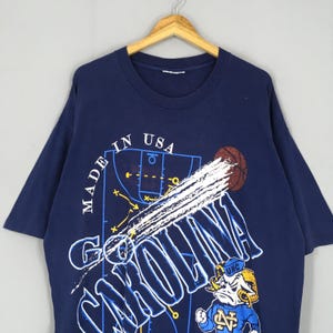 May include: Navy blue t-shirt featuring a basketball court design with the text "CAROLINA" in white. The shirt includes a basketball graphic and a cartoon ram mascot. The text "MADE IN USA" is also visible.