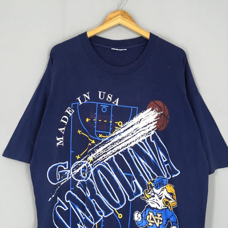 May include: Navy blue t-shirt featuring a basketball court design with the text "CAROLINA" in white. The shirt includes a basketball graphic and a cartoon ram mascot. The text "MADE IN USA" is also visible.