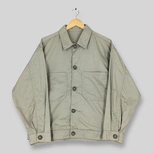 May include: A light olive green jacket with a button-up front, two chest pockets, and buttoned cuffs. The jacket is hanging on a wooden hanger against a light gray background. The jacket has a classic, casual style.