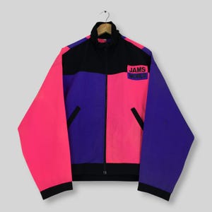May include: A color-blocked zip-up jacket featuring a black upper chest and collar, with vibrant pink and purple panels. The jacket includes a black front zipper, two side pockets, and a rectangular patch on the chest with the text JAMS WORLD.