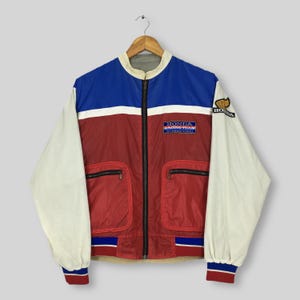 May include: A vintage Honda Racing Team International jacket. The jacket features a color block design with blue, red, and white panels. It has a zip-up front, two front pockets, and the Honda logo on the chest and sleeve. The jacket is hanging on a wooden hanger.
