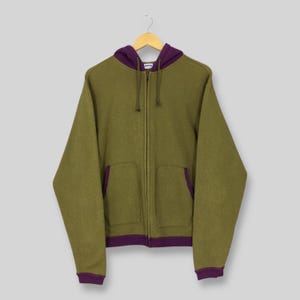 Peut inclure: Un sweat à capuche vert olive zippé avec une doublure de capuche, une bordure de poche et des poignets violets. Le sweat à capuche a deux poches avant et une fermeture éclair intégrale. Suspendu à un cintre en bois sur fond gris clair.