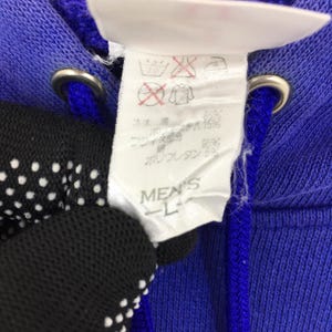 May include: Close-up of a blue hoodie with a white tag. The tag has washing instructions and the text "MEN'S L". The hoodie has a drawstring and metal eyelets.