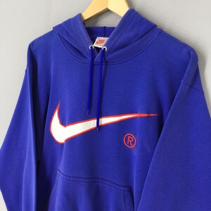 May include: A royal blue hooded sweatshirt featuring a large white Nike swoosh logo with an orange outline. The sweatshirt has a kangaroo pocket and a drawstring hood. The Nike logo and registered trademark symbol are visible.