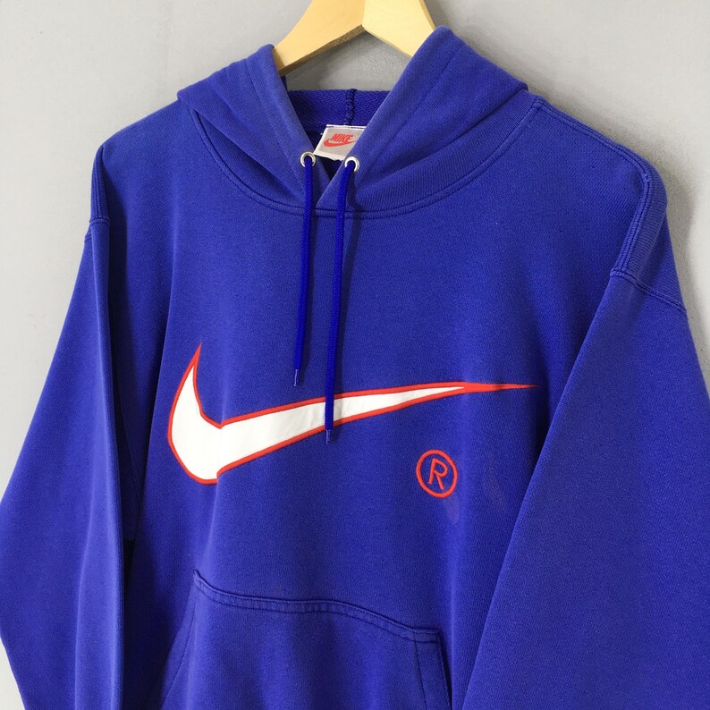 May include: A royal blue hooded sweatshirt featuring a large white Nike swoosh logo with an orange outline. The sweatshirt has a kangaroo pocket and a drawstring hood. The Nike logo and registered trademark symbol are visible.