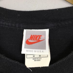 Puede incluir: Primer plano del cuello de una camiseta negra con una etiqueta Nike. La etiqueta presenta el logotipo de Nike en rojo y blanco, con el texto "MADE IN U.S.A." e informaci&oacute;n de tallas. La camiseta es talla M.