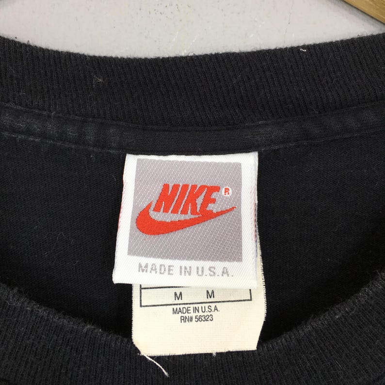 Puede incluir: Primer plano del cuello de una camiseta negra con una etiqueta Nike. La etiqueta presenta el logotipo de Nike en rojo y blanco, con el texto "MADE IN U.S.A." e informaci&oacute;n de tallas. La camiseta es talla M.