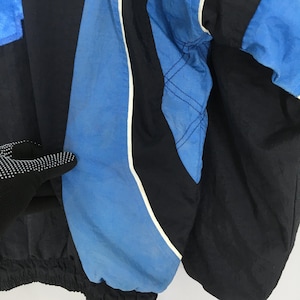 May include: Close-up of a black and blue vintage jacket. The jacket features blue panels with white trim and black sections. The jacket has a shiny, nylon-like texture. The sleeve is visible, with a black cuff and a blue panel.