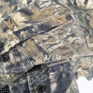 May include: Close-up of a camouflage button-down shirt. The shirt features a pattern of green, brown, and gray, with visible buttons and a sleeve cuff. The fabric shows signs of wear and tear, with small holes near the cuff.