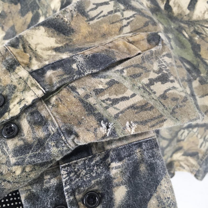 May include: Close-up of a camouflage button-down shirt. The shirt features a pattern of green, brown, and gray, with visible buttons and a sleeve cuff. The fabric shows signs of wear and tear, with small holes near the cuff.