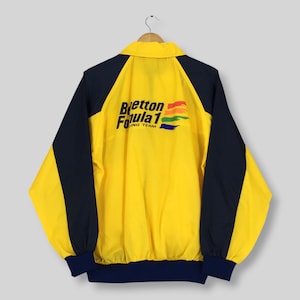 Puede incluir: Una chaqueta cortavientos amarilla y azul marino con cuello y mangas largas. La parte posterior muestra el texto Benetton Formula 1 Racing Team en letras negras, acompañado de un gráfico de cuatro trazos de pincel horizontales en rojo, naranja, verde y azul.