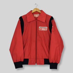 May include: A red zip-up jacket with black panels on the sleeves and waist. The jacket features a white patch on the chest with the text Racing Development TRD and a matching label on the inside collar. It has a pointed collar and long sleeves.