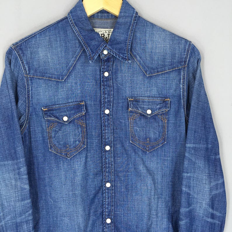 May include: A medium-wash denim shirt with a Western-style yoke and two chest pockets. The shirt has white buttons down the front and on the pockets. The pockets have decorative stitching. The shirt is hanging on a wooden hanger.