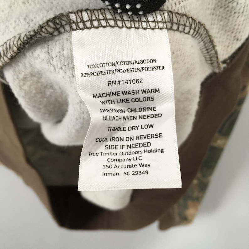 May include: A close-up of a clothing label with care instructions. The white label has black text detailing fabric composition, RN number, and washing instructions. The garment has a camouflage pattern.