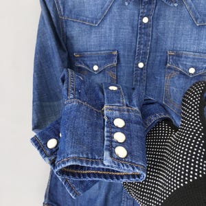 May include: A blue denim shirt with white buttons and two front pockets. The shirt has long sleeves with buttoned cuffs. The fabric is a medium wash denim.